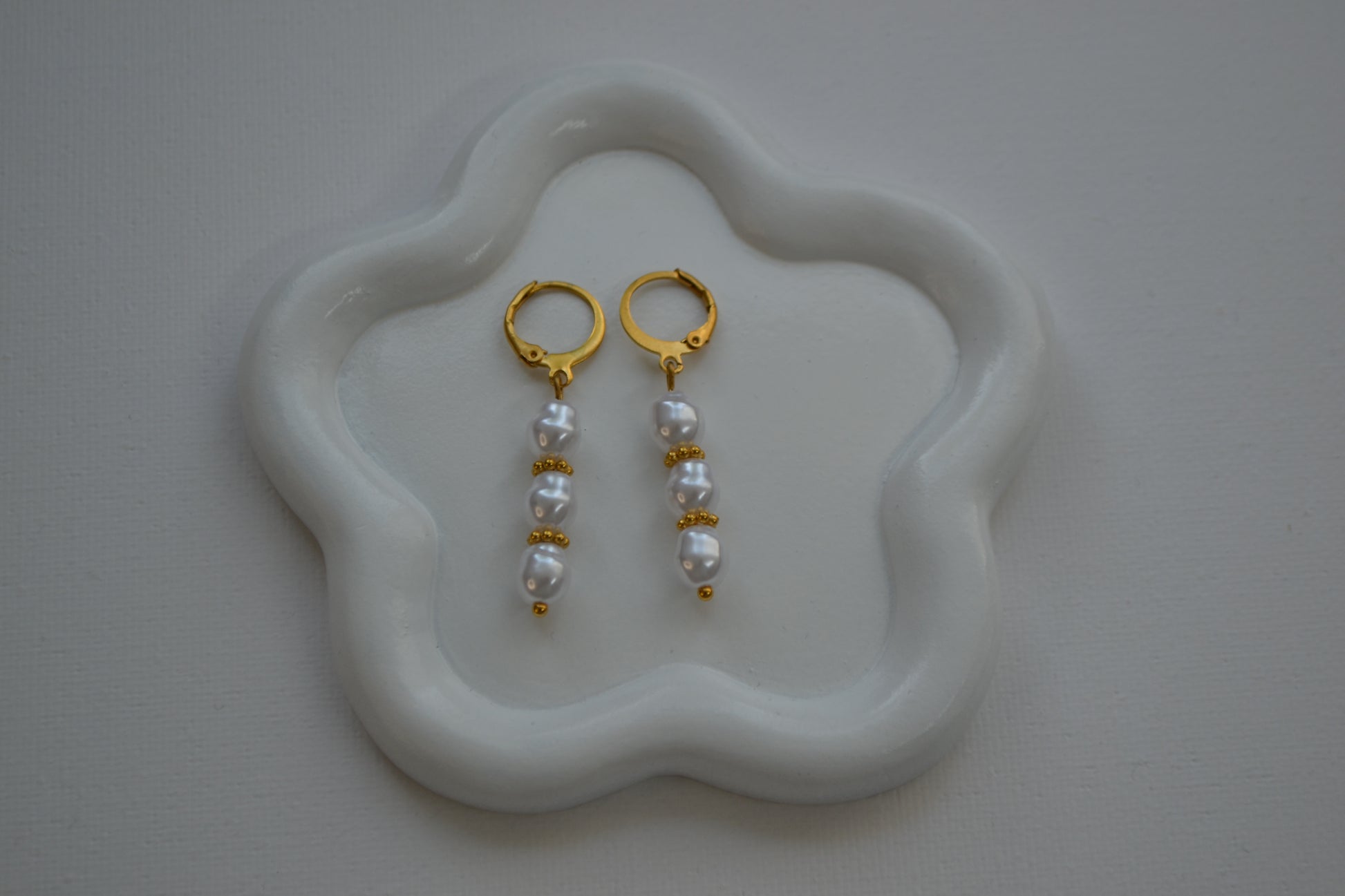 Oorbellen met een gouden clip en drie parels met gouden spacers ertussen. De oorbellen liggen op een wit bloem bordje met een witte achtergrond.