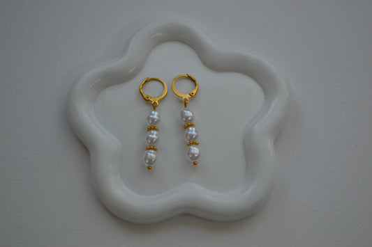 Oorbellen met een gouden clip en drie parels met gouden spacers ertussen. De oorbellen liggen op een wit bloem bordje met een witte achtergrond.