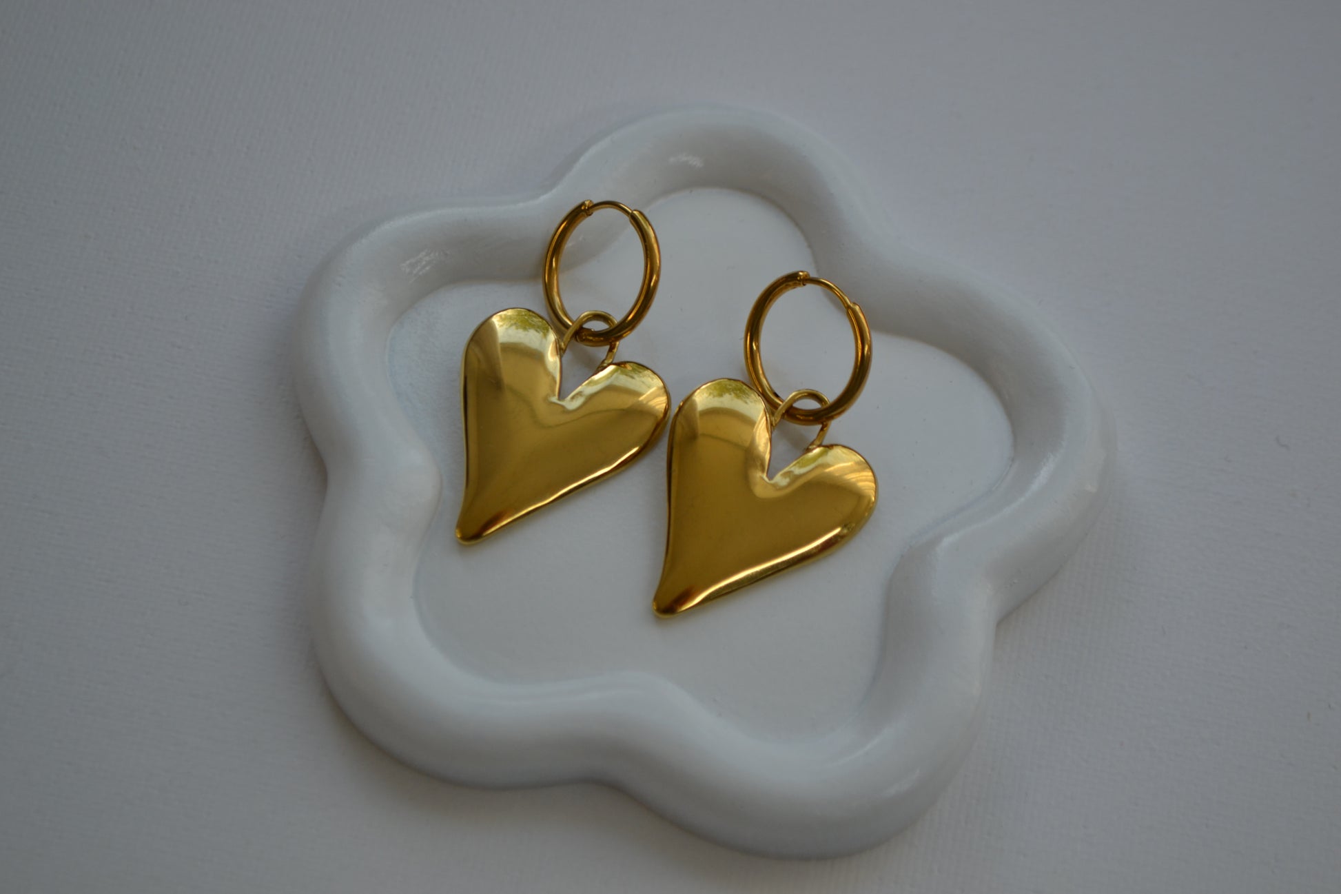 Gouden oorbellen met een creool en een grote bedel in de vorm van een hart. De oorbellen liggen op een wit bordje in de vorm van een bloem op een witte achtergrond.