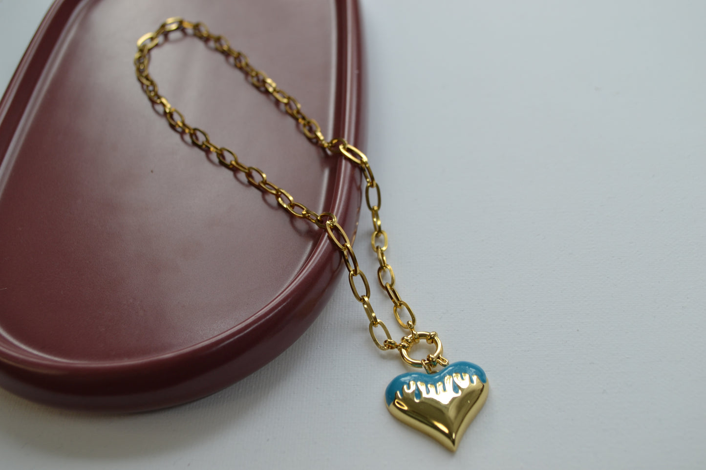 Gouden ketting met een bedel met goud en blauw op een schaaltje van donker rode kleur en een witte achtergrond.