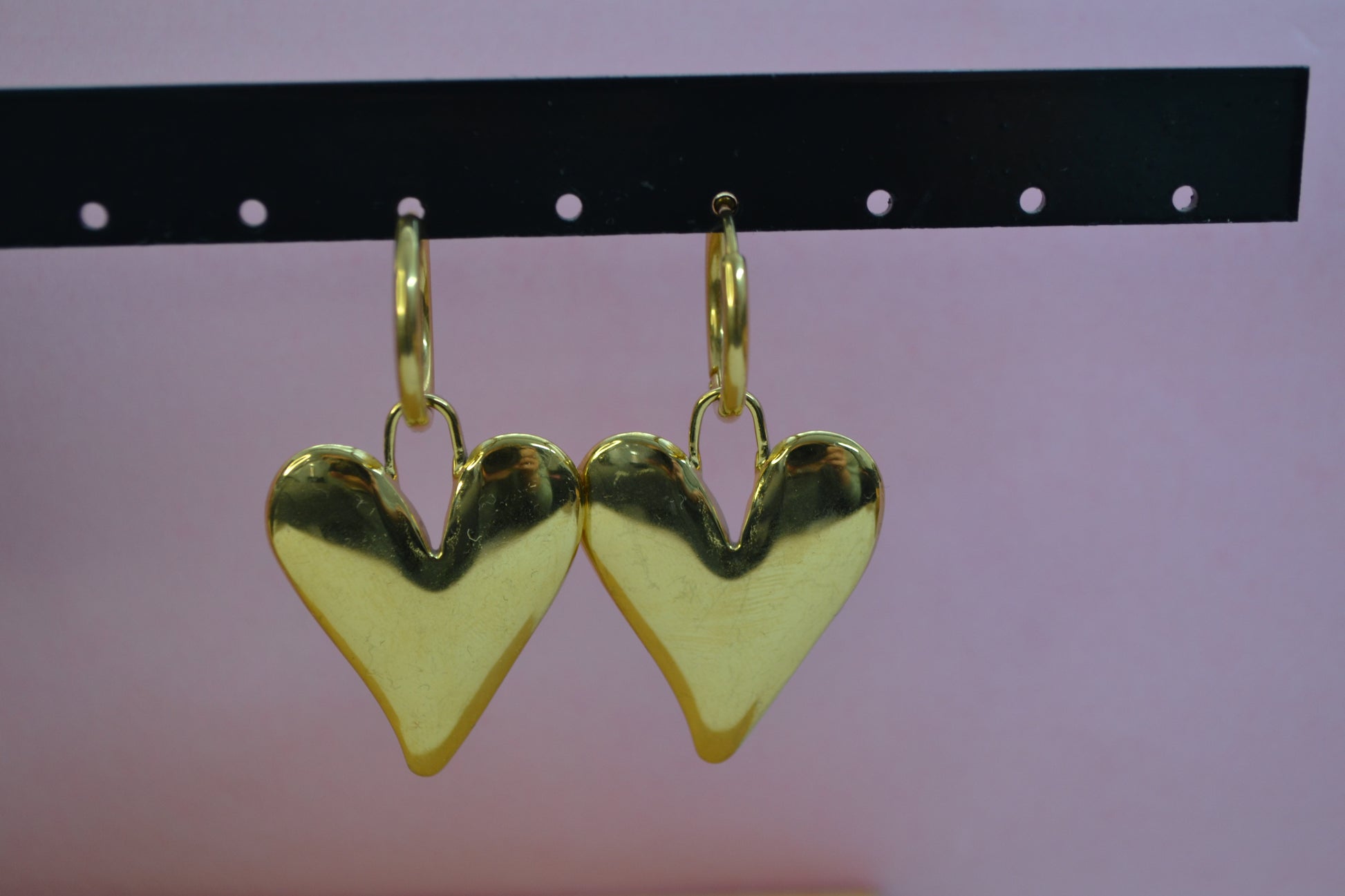 Gouden oorbellen met een creool en een grote bedel in de vorm van een hart. De oorbellen hangen op een rekje en de achtergrond is roze.