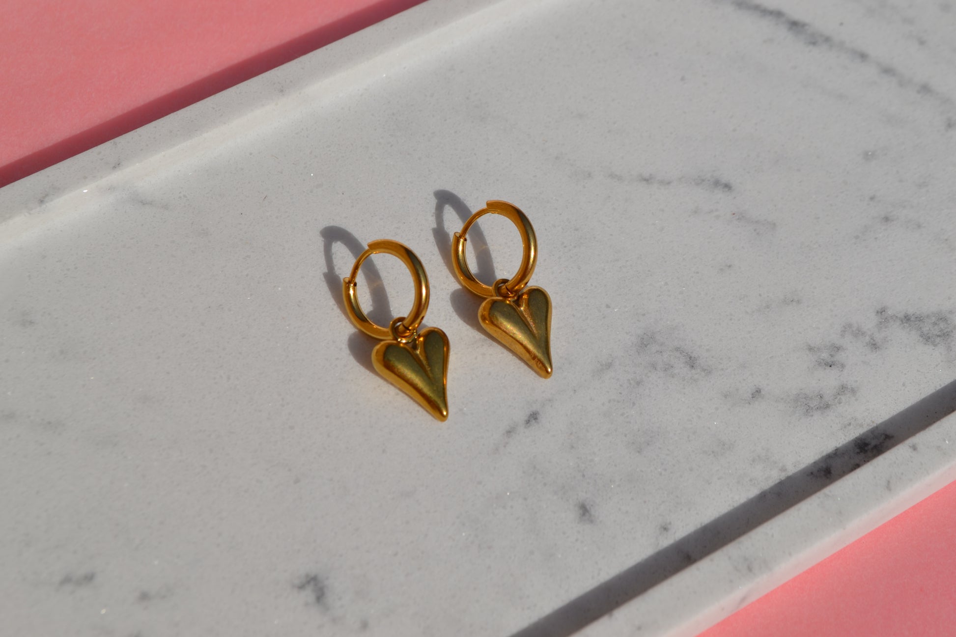 Oorbellen met gouden creoolen en een gouden bedel in de vorm van een hartje. De oorbellen liggen op een marmeren wit schaaltje op een roze ondergrond.