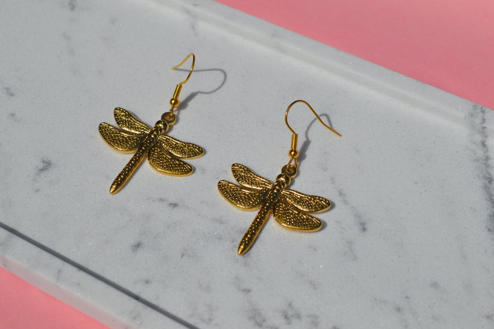 Gouden oorbellen met een haakje en een bedel in de vorm van een libelle op een wit marmeren plankje met een roze achtergrond.