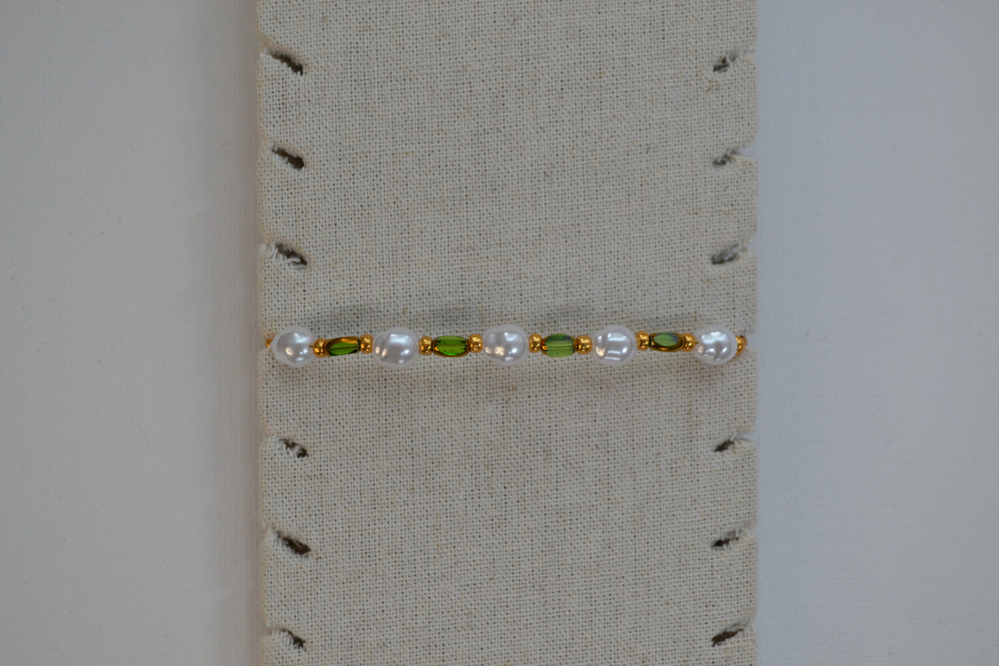 Een armband met een gouden sluiting en groene, gouden en parel kralen. De armband zit op een beige armband stand.