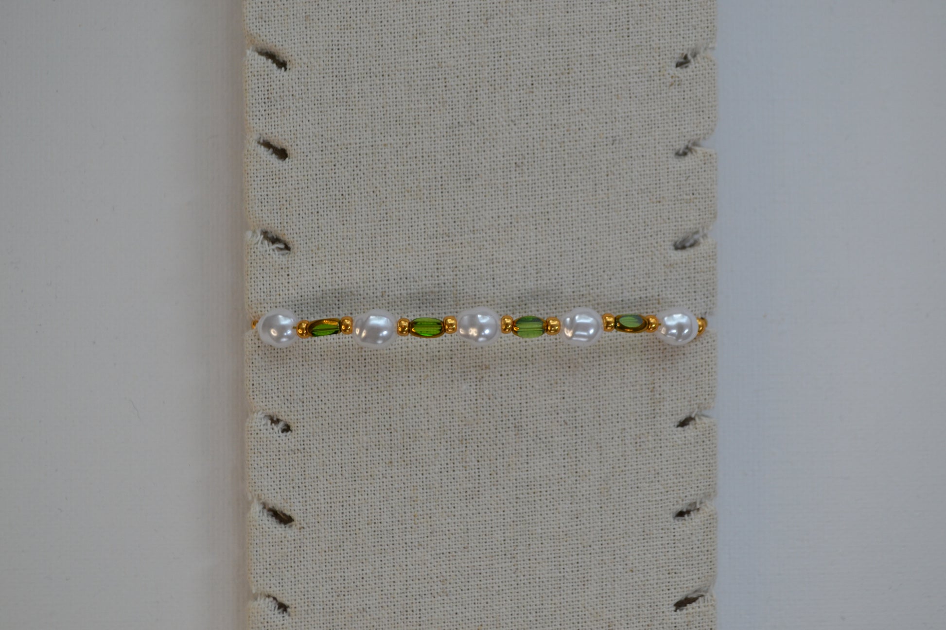Een armband met een gouden sluiting en groene, gouden en parel kralen. De armband zit op een beige armband stand.