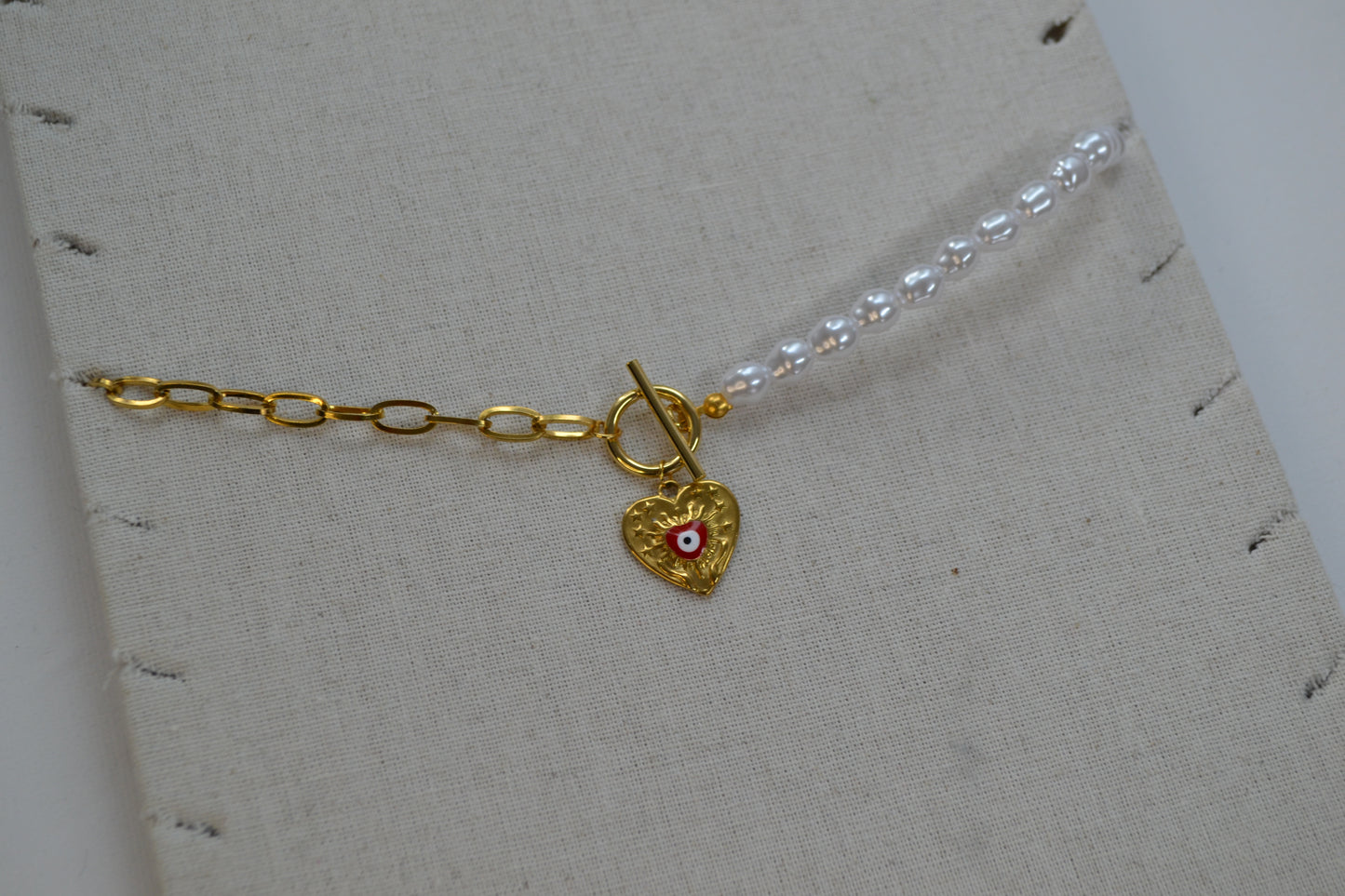 Een ketting met aan de ene kant een gouden ketting en aan de andere kant een parelketting. De ketting heeft een kapitelsluiting en een gouden bedel in de vorm van een hart met een rood oogje. De ketting zit op een beige ketting stand.