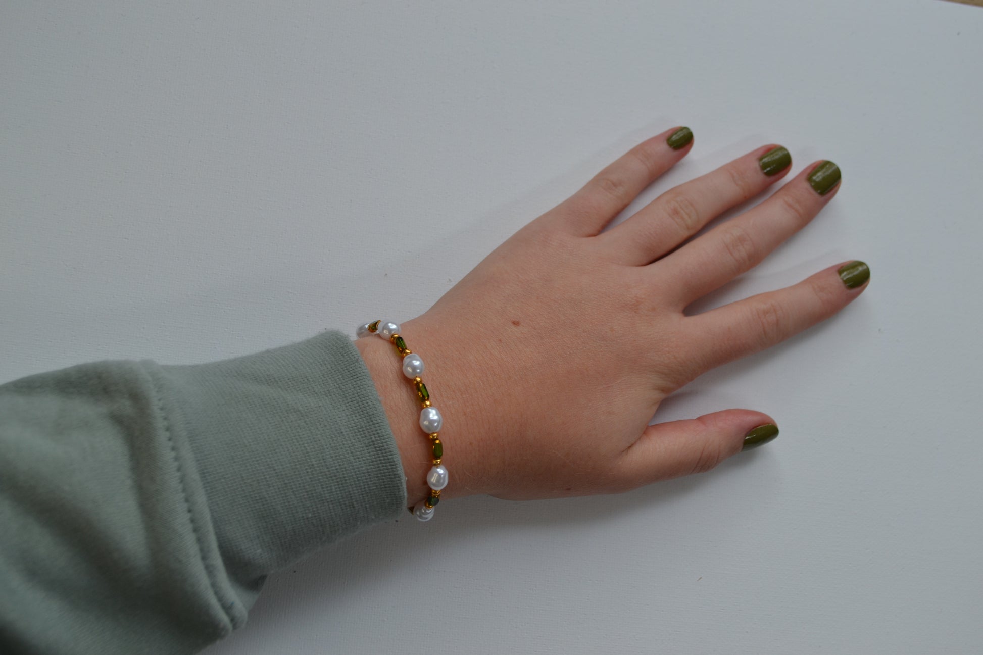 Een armband met een gouden sluiting en groene, gouden en parel kralen. De armband wordt om de pols gedragen en ligt op een witte ondergrond.