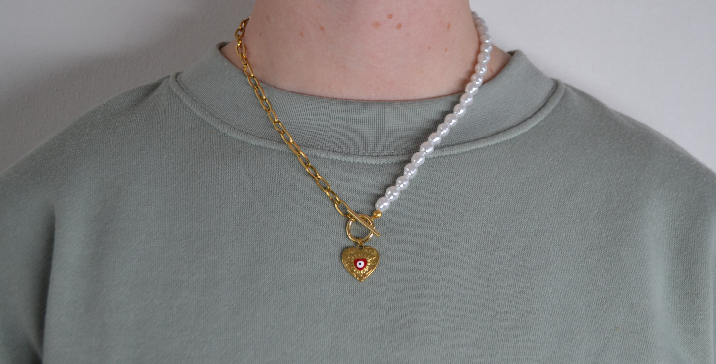 Een ketting met aan de ene kant een gouden ketting en aan de andere kant een parelketting. De ketting heeft een kapitelsluiting en een gouden bedel in de vorm van een hart met een rood oogje. Een vrouw draagt de ketting.
