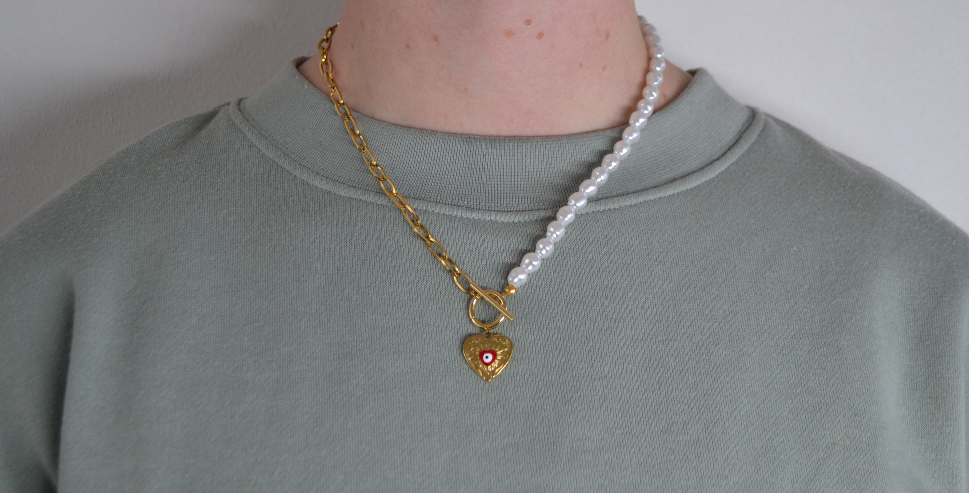 Een ketting met aan de ene kant een gouden ketting en aan de andere kant een parelketting. De ketting heeft een kapitelsluiting en een gouden bedel in de vorm van een hart met een rood oogje. Een vrouw draagt de ketting.
