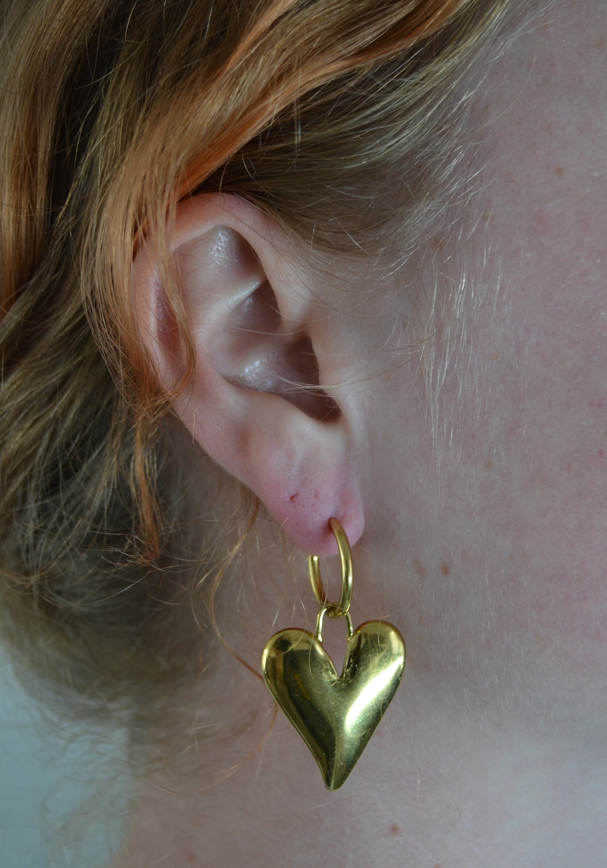 Gouden oorbellen met een creool en een grote bedel in de vorm van een hart. Een vrouw draagt de oorbellen.