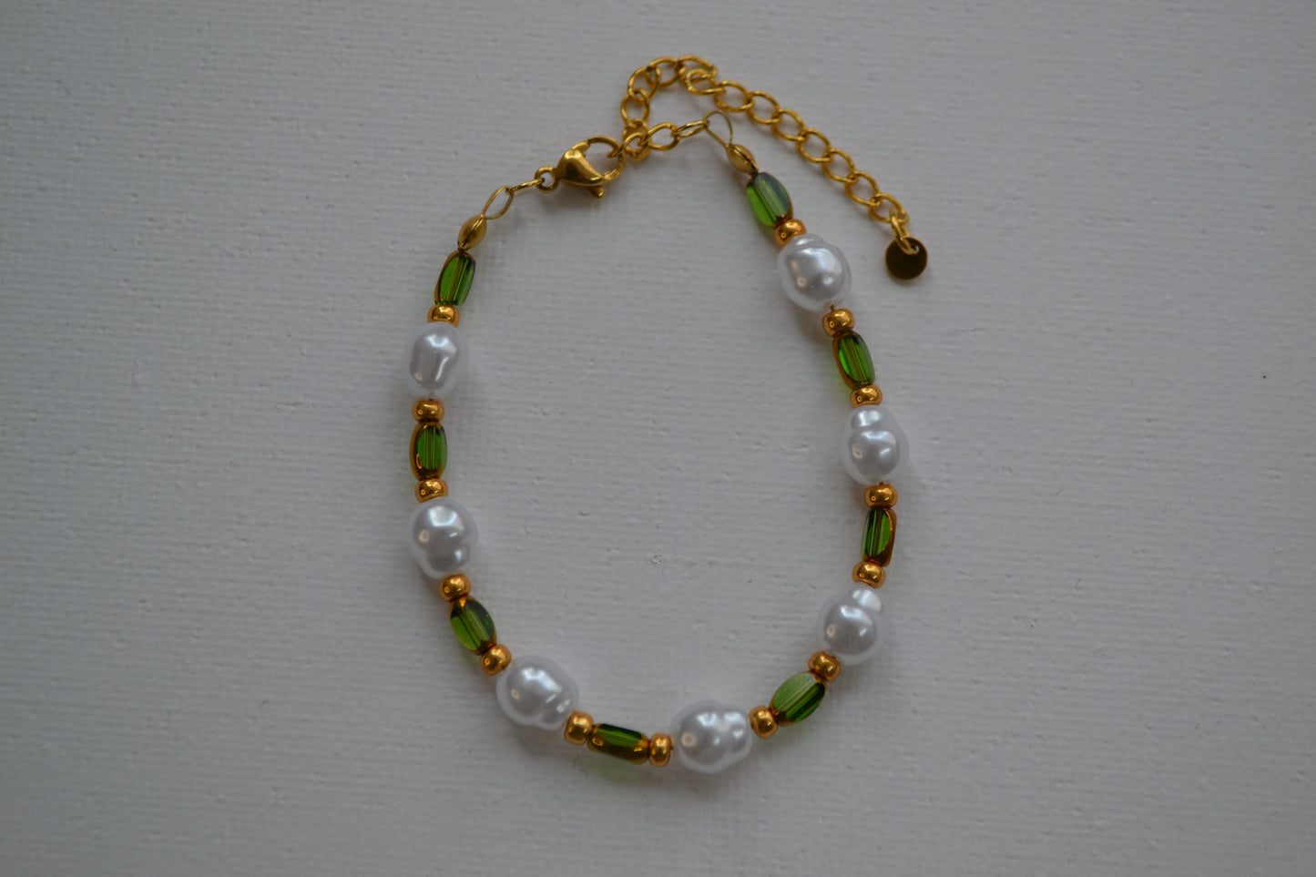 Een armband met een gouden sluiting en groene, gouden en parel kralen. De armband ligt op een witte ondergrond.