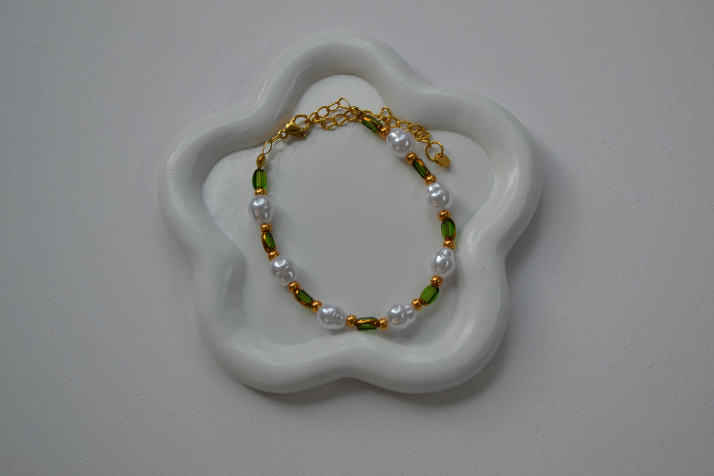 Een armband met een gouden sluiting en groene, gouden en parel kralen. De armband ligt op een wit schaaltje in de vorm van een bloem. Het schaaltje staat op een witte ondergrond.