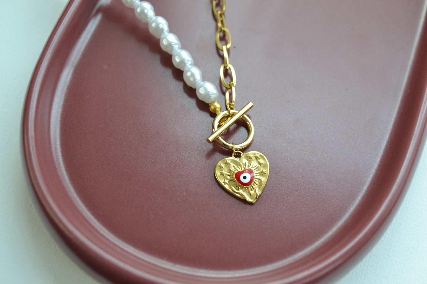 Een ketting met aan de ene kant een gouden ketting en aan de andere kant een parelketting. De ketting heeft een kapitelsluiting en een gouden bedel in de vorm van een hart met een rood oogje. De foto is ingezoomed op de bedel en deze ligt op een bruin schaaltje.