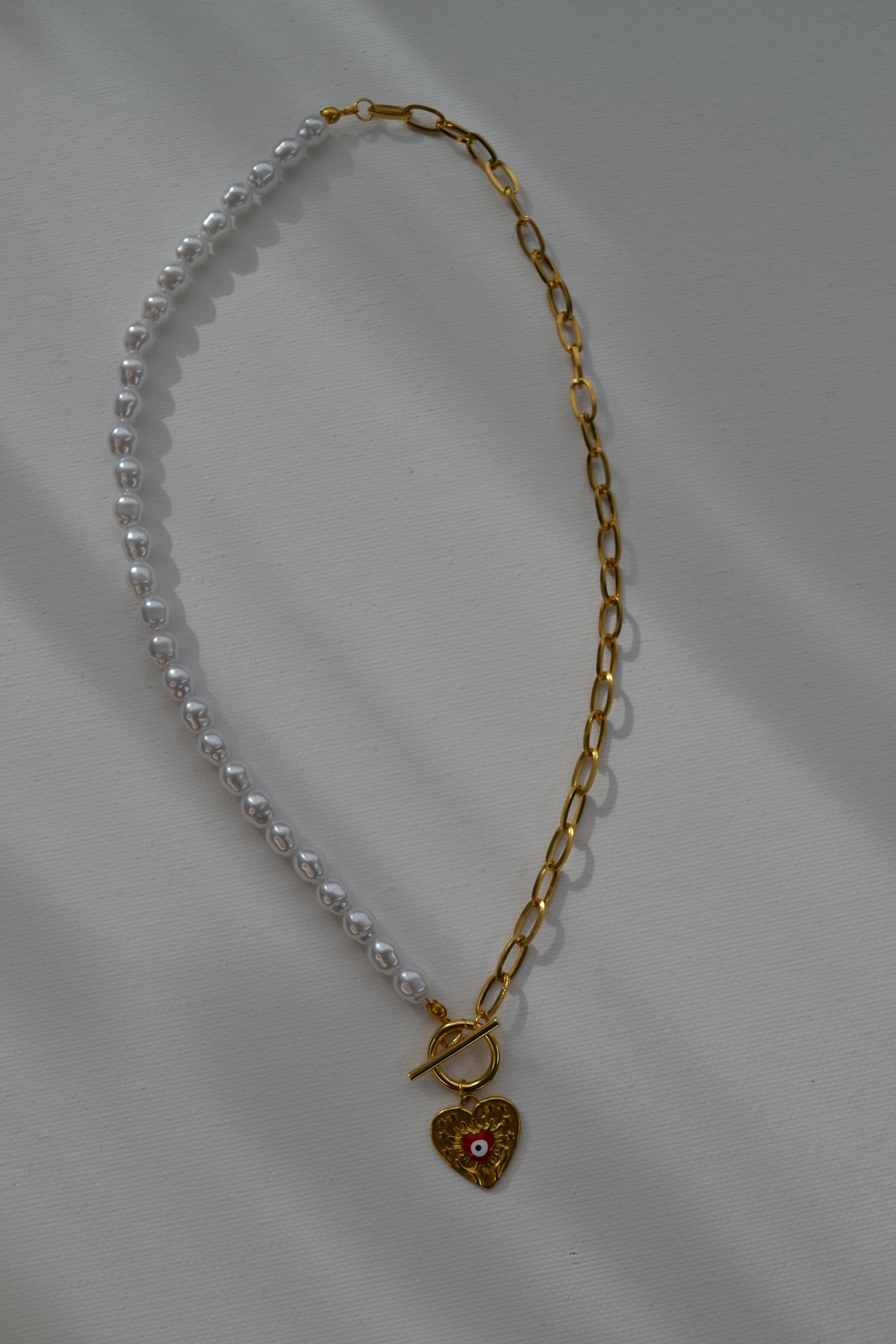 Een ketting met aan de ene kant een gouden ketting en aan de andere kant een parelketting. De ketting heeft een kapitelsluiting en een gouden bedel in de vorm van een hart met een rood oogje. De ketting ligt op een witte achtergrond.