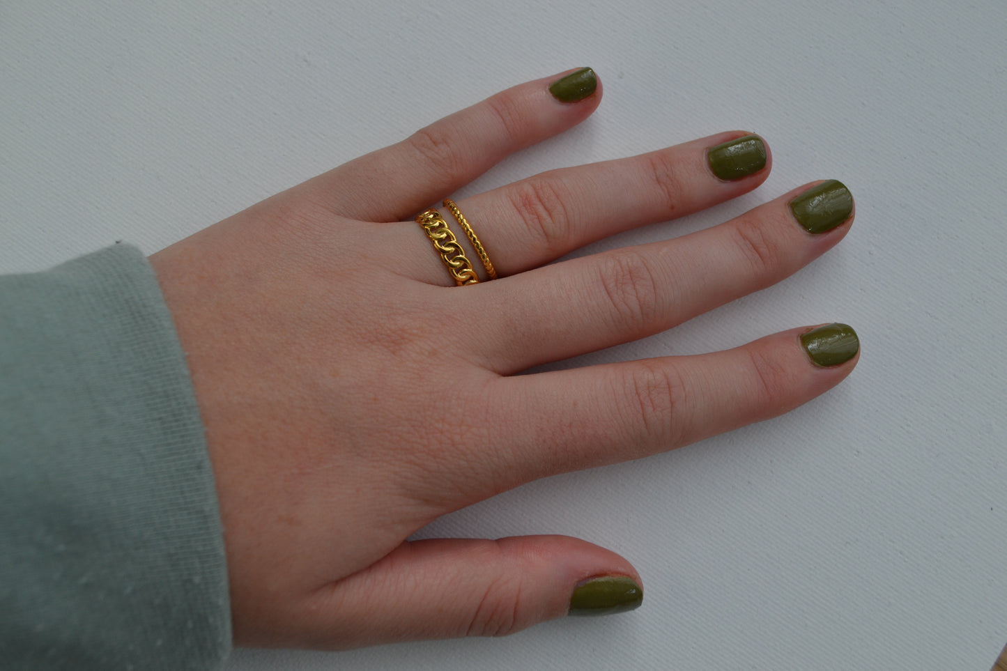Gouden ring met een dubbele band op een hand waarvan de nagels groen zijn gelakt en op een witte achtergrond.