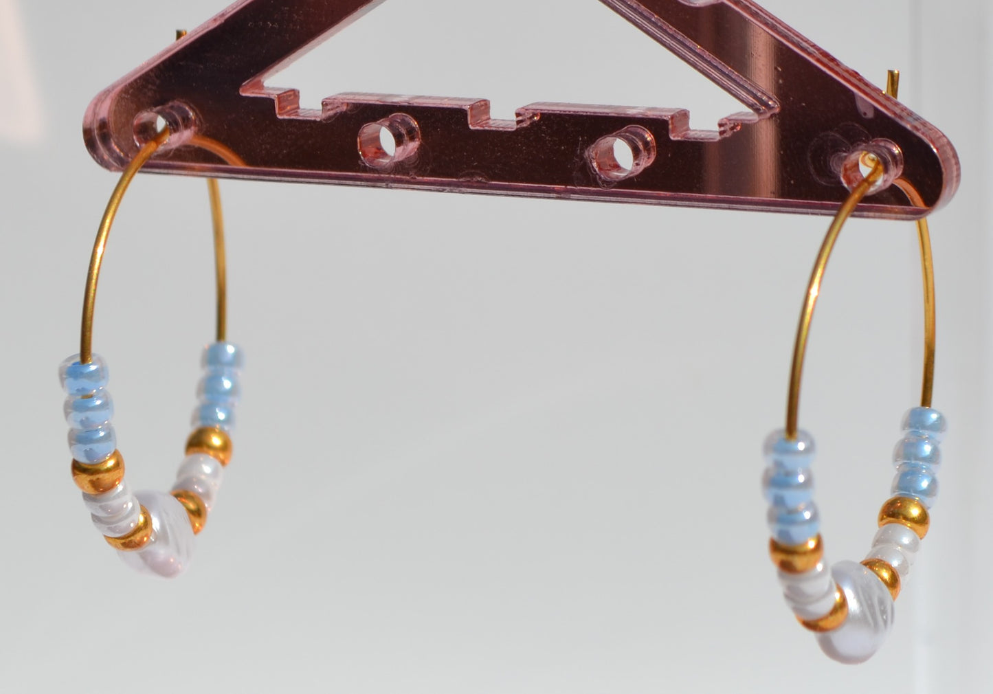 Gouden hoepeloorbellen met witte, gouden en blauwe kralen en een parel. De oorbellen hangen aan een roze rekje.