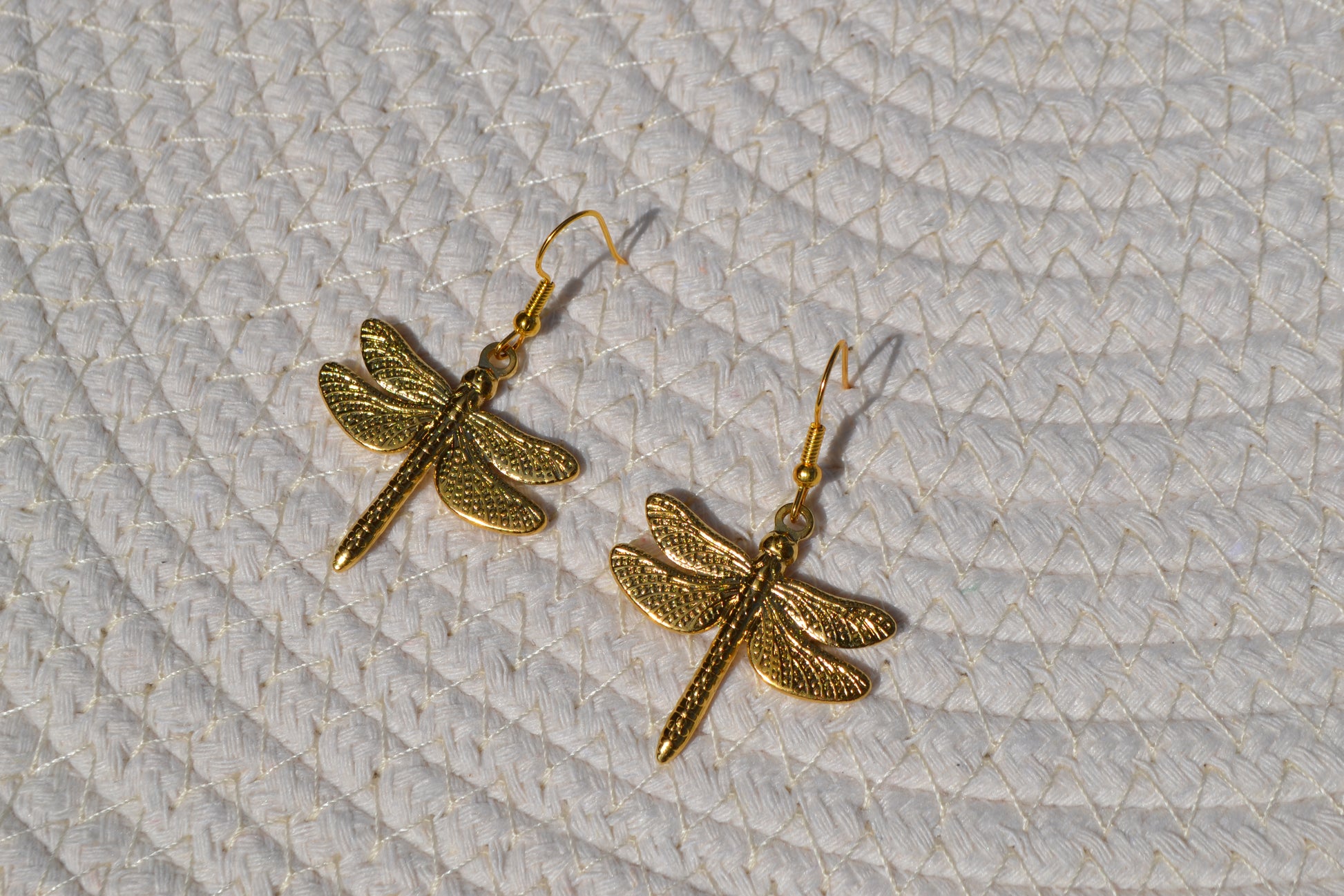 Gouden oorbellen met een haakje en een bedel in de vorm van een libelle op een wit matje.