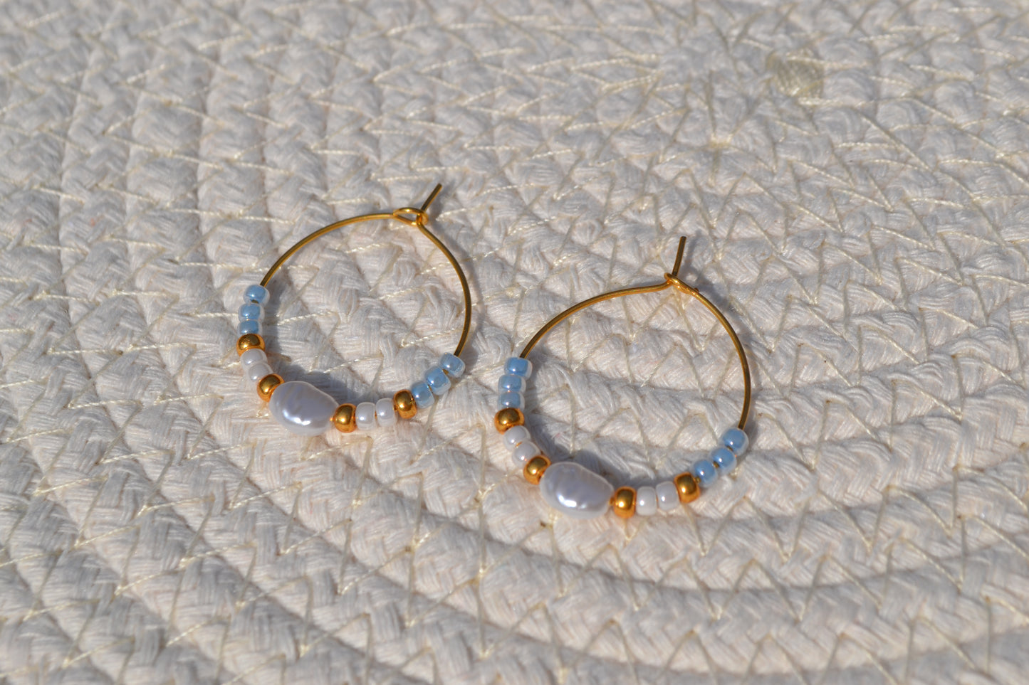 Gouden hoepeloorbellen met witte, gouden en blauwe kralen en een parel. De oorbellen liggen op een wit matje.