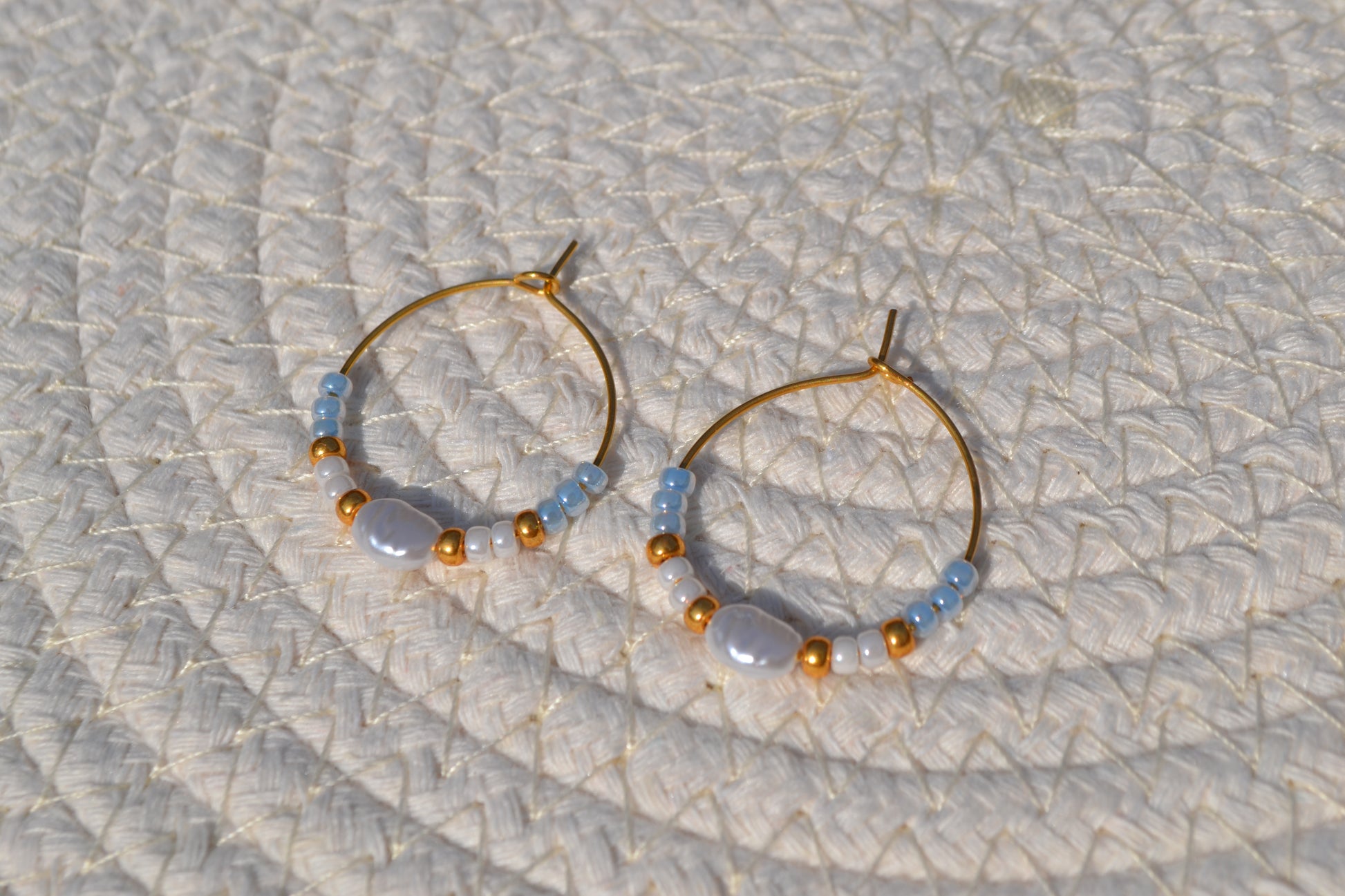 Gouden hoepeloorbellen met witte, gouden en blauwe kralen en een parel. De oorbellen liggen op een wit matje.