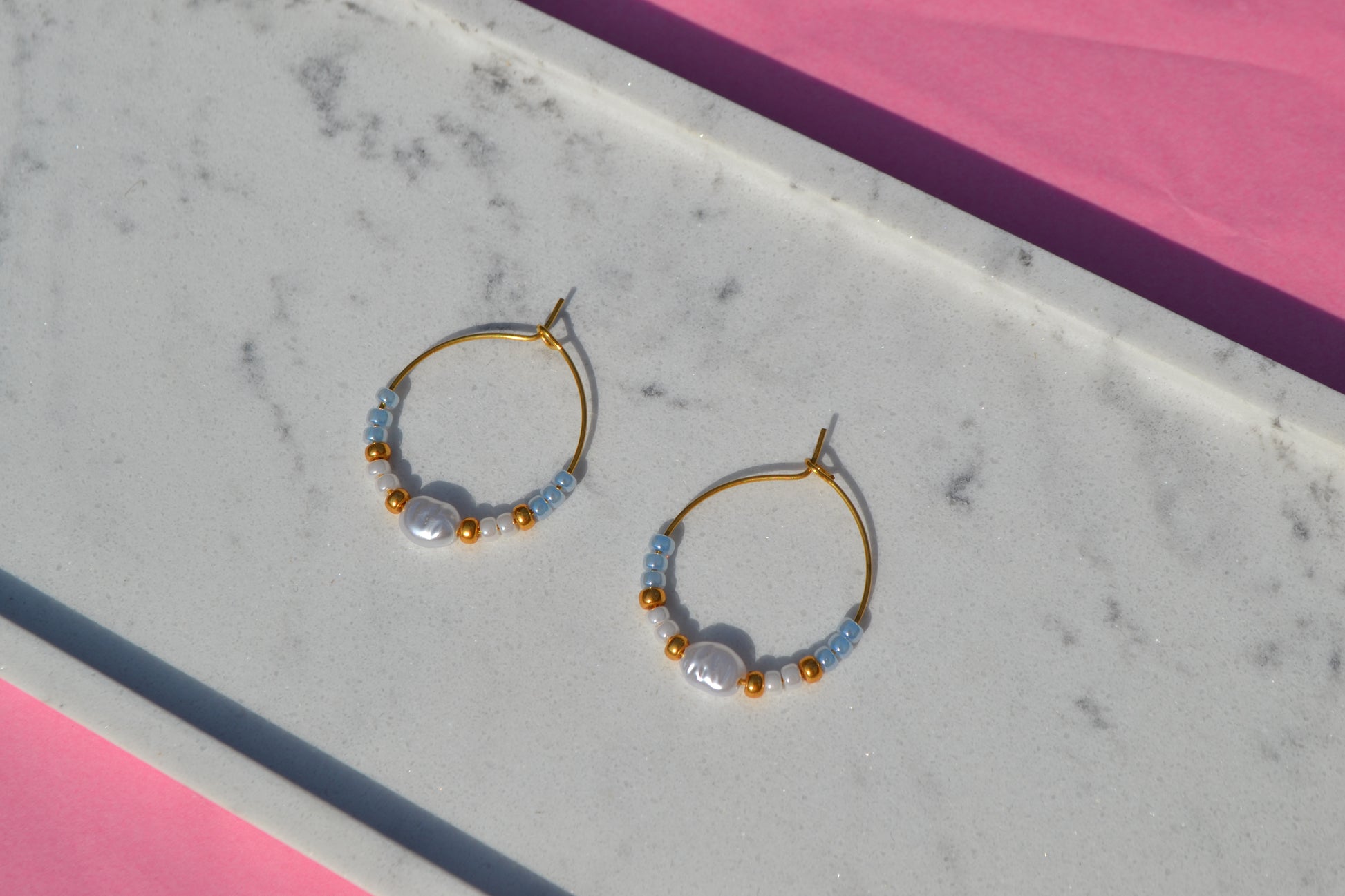 Gouden hoepeloorbellen met witte, gouden en blauwe kralen en een parel. De oorbellen liggen op een wit marmeren plankje op een roze achtergrond.