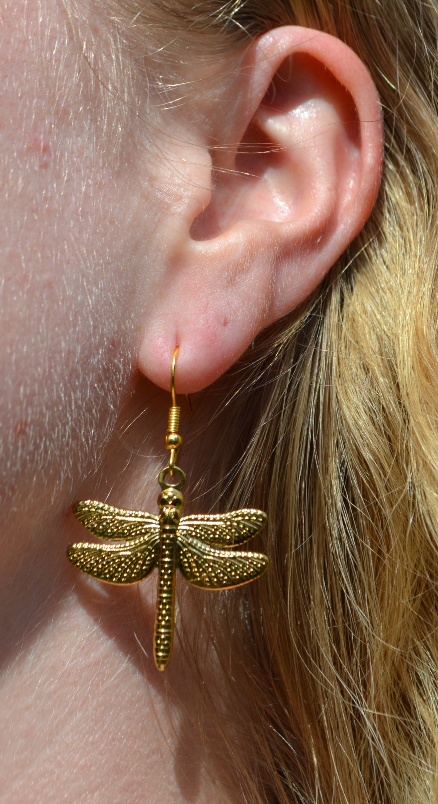 Gouden oorbellen met een haakje en een bedel in de vorm van een libelle. De oorbellen hangen in de oren van een vrouw.